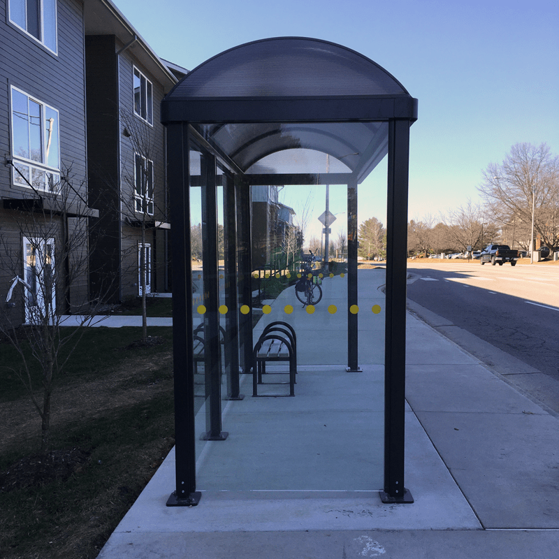 A bus shelter with all glass sides is seen from the left side with bench seating inside.