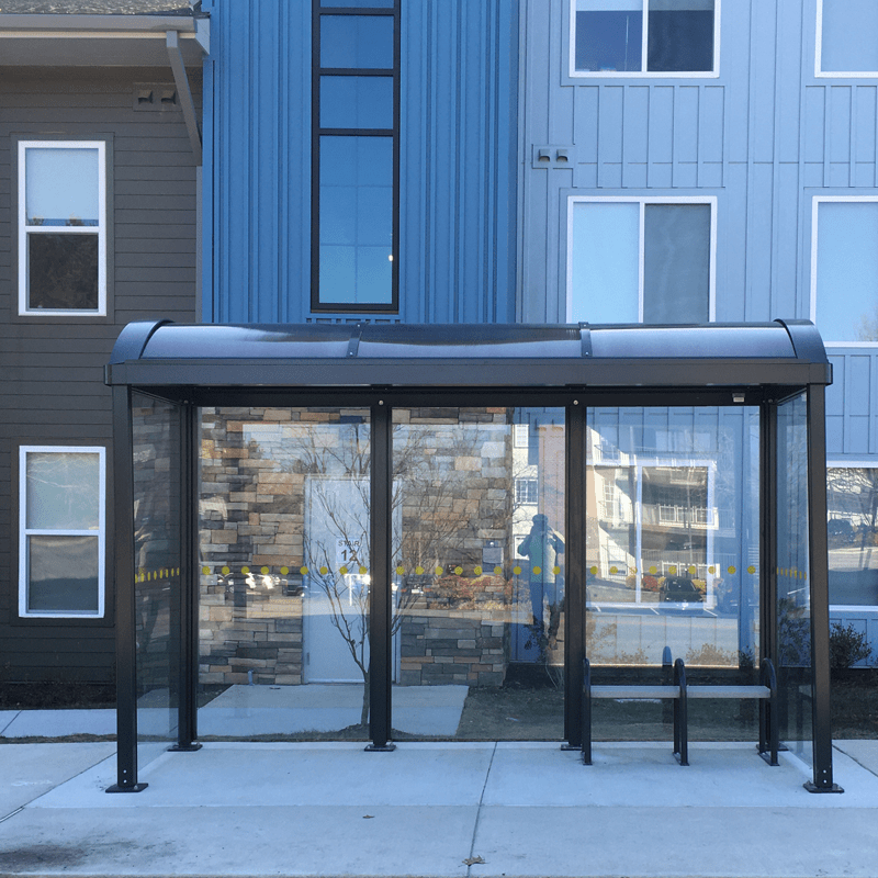 A bus shelter is seen from the front with bench seating inside the shelter and glass siding.