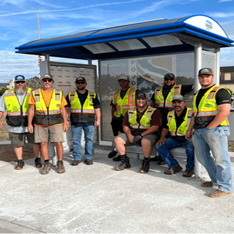 A group of men with safety vests are seen sitting and standing around a bus shelter.
