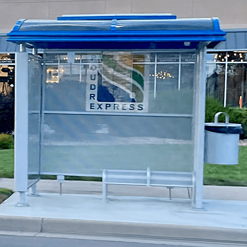 A bus shelter is seen from the front with a left panel advertising display, a trash receptacle attached on the right and bench seating inside the shelter.