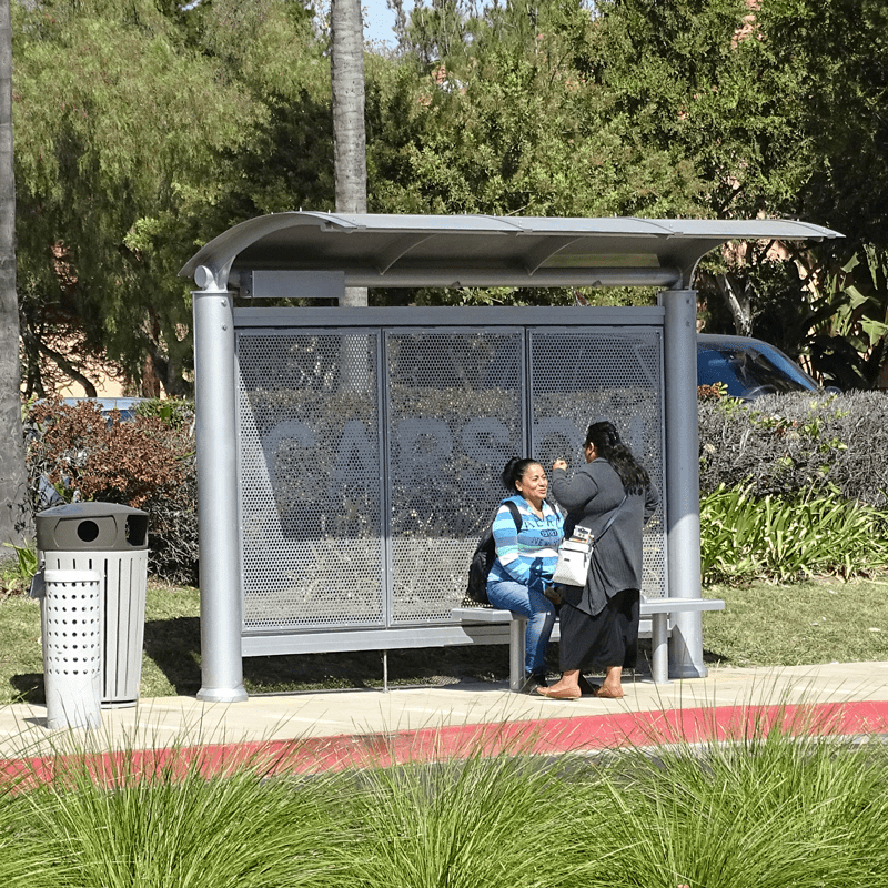 Front stepped back view of a Signa Sunset custom bus shelter. Two passengers await the bus while one sits on the bench