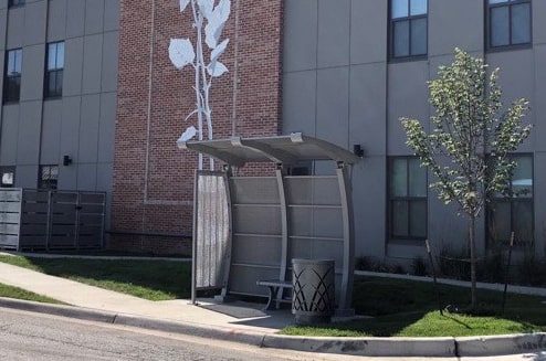 Small bus shelter with small bench and trash receptacle outside a building on the corner of the street