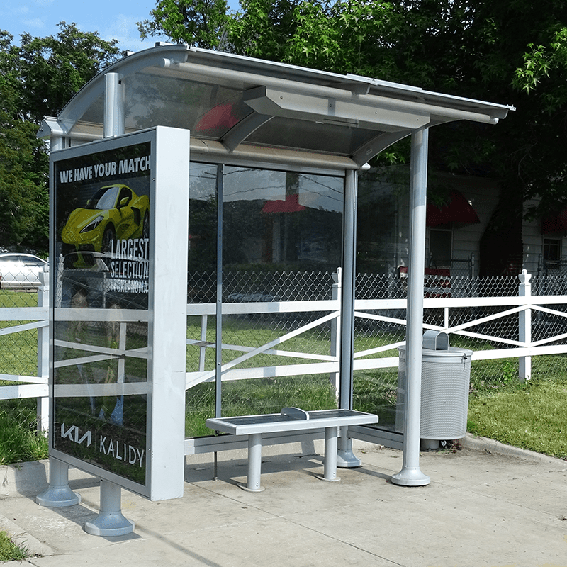Left facing bus shelter includes a bench for seating and a double sides advertising display on the left, trash can on the right