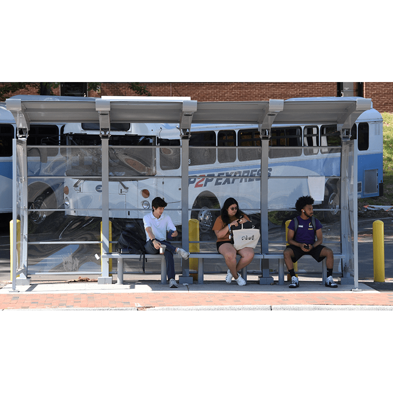 Forward view of a Signa Crescent bus shelter in light blue with seating for three passengers as they wait for their bus with a bus in the background of the shelter