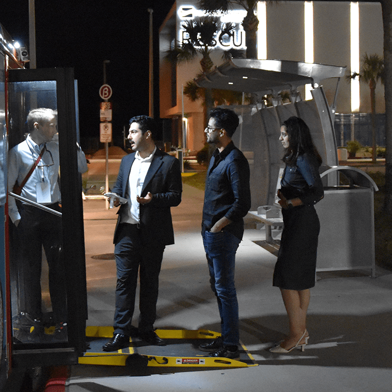Riders talk to one another as they wait to board the bus through the bus ramp with a bus shelter in the near distance
