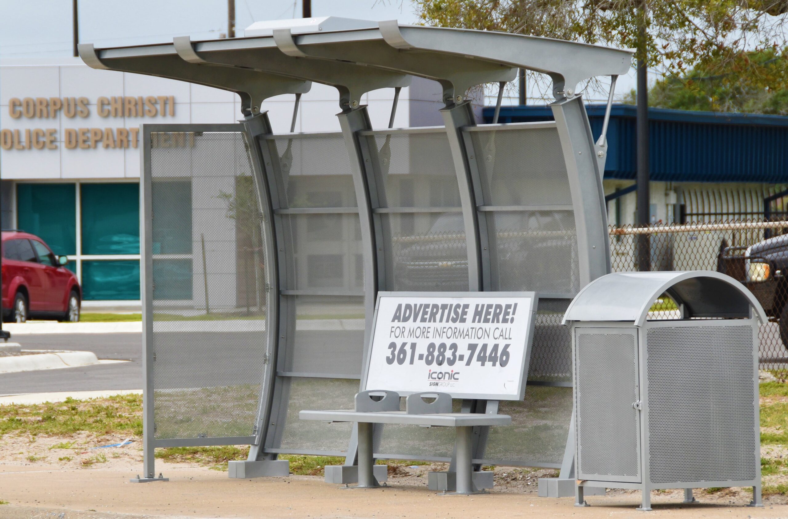 Left side view of a bus shelter that offers bench seating with advertising on the back rest of the bench and a trash receptacle on the left side