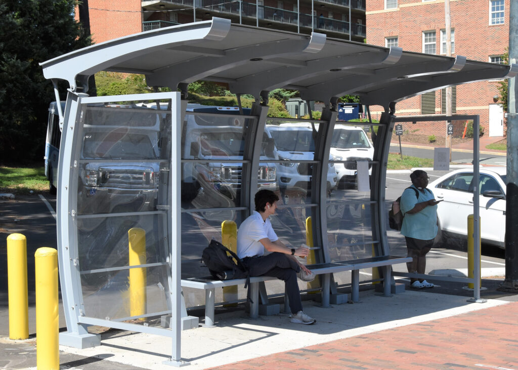 Two passengers wait in the shade under a bush shelter designed with a bench or standing room
