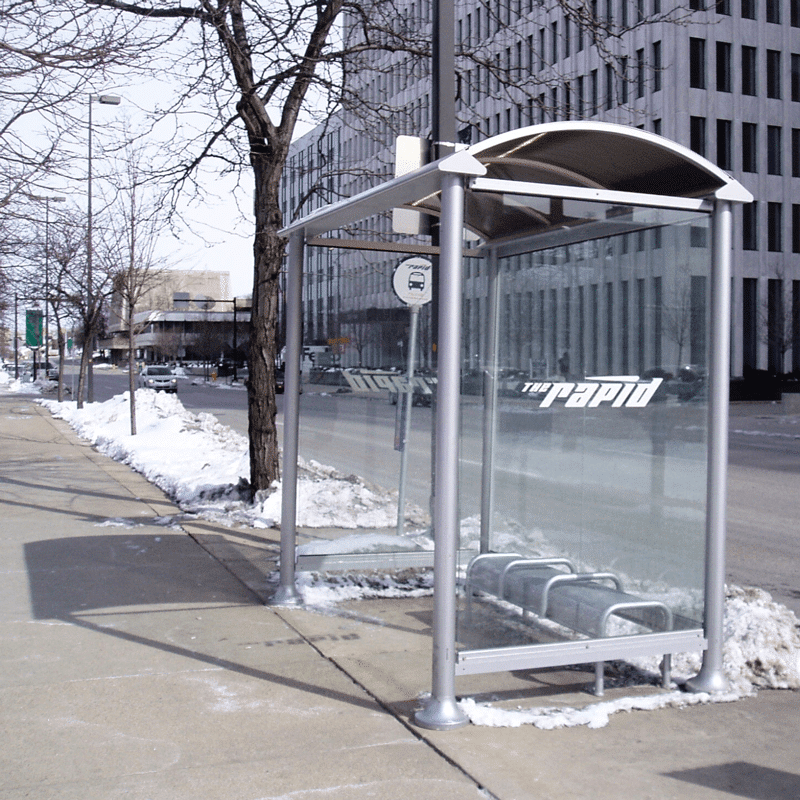 A bus shelter for The Rapid in Grand Rapids is seen from the left side with glass siding and a bench for three.