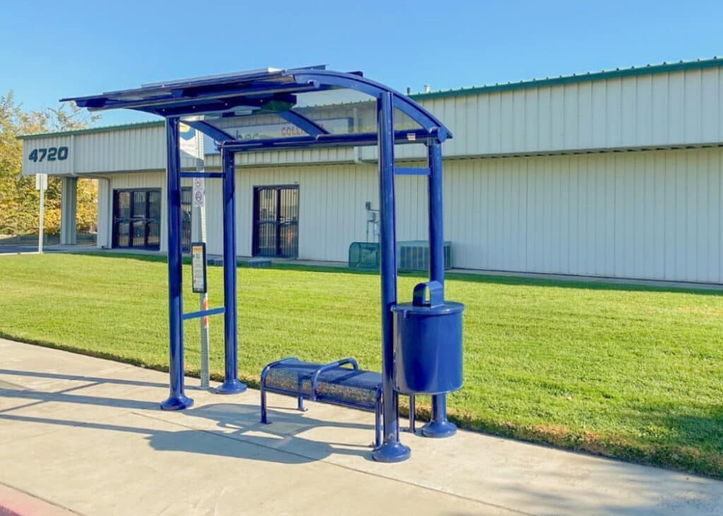 A blue bus shelter is seen head on outside a building with a trash receptacle attached and bench seating for two