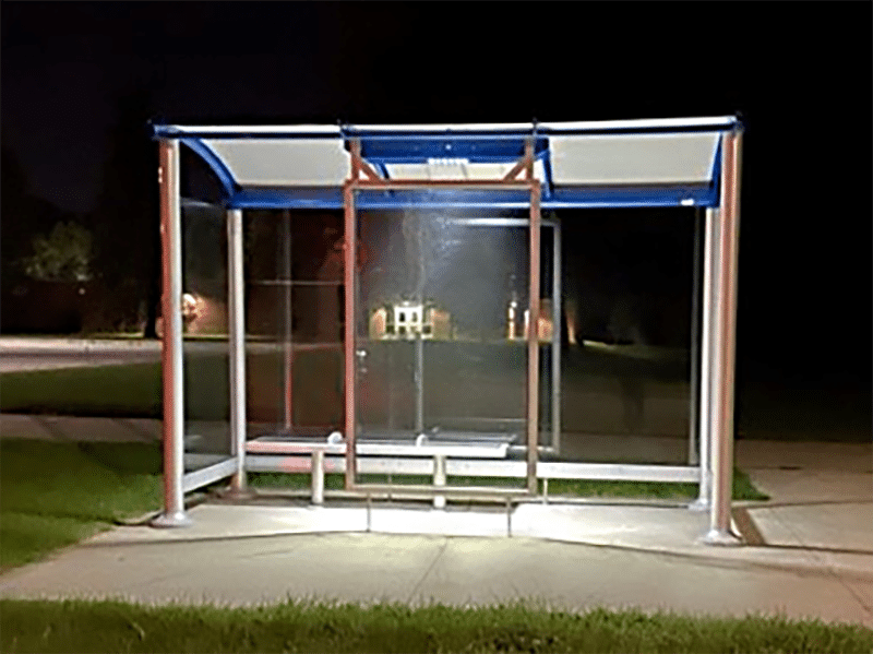 A Port Huron bus shelter is seen as night from the front with lights on showing the three seat bench