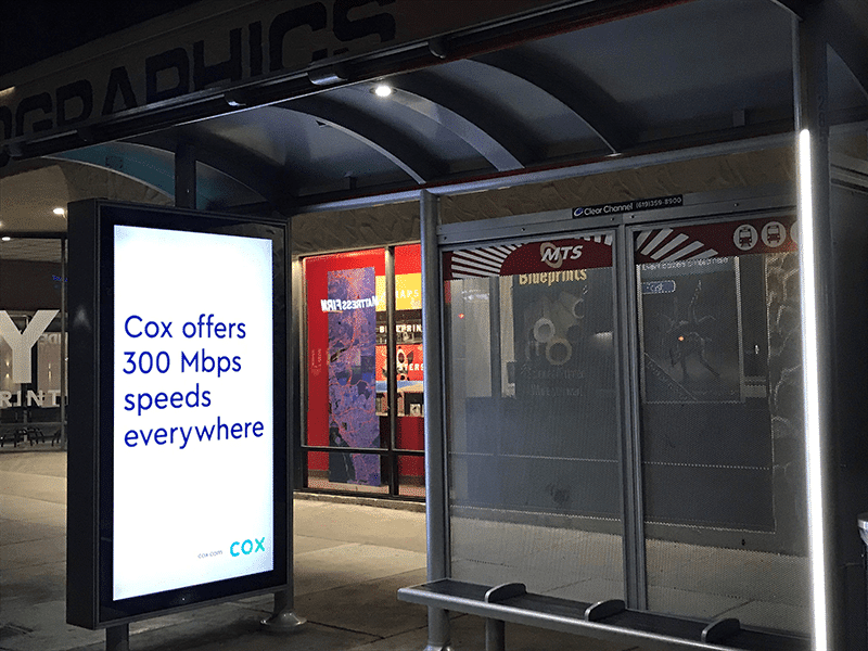 A left side view of a bus shelter at night with an lit advertising display and bench seating