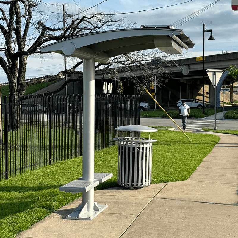 An eco-shade bus shelter is seen from the left side with seats on either side of the pole shade and a trash receptacle next to the shelter.