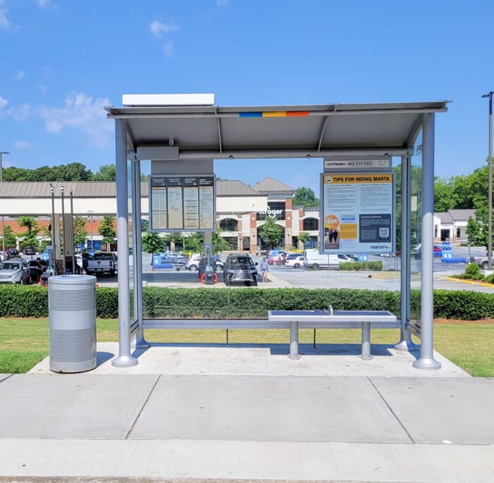 A bus shelter is seen from the front with bench seating for two on the right and a trash receptacle on the left. Also equipped with rider signage on either side.