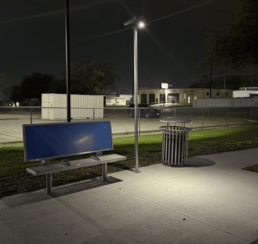 A bus stop is seen at night from the left side with a three seat bench with backrest advertising, a lit light post lit and a trash receptable to the right.