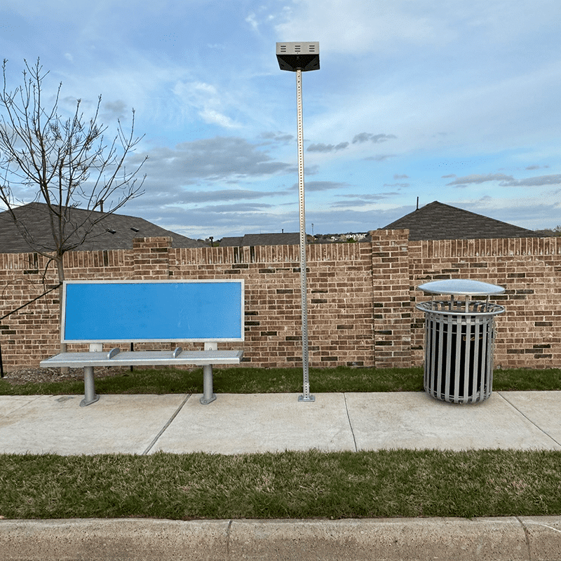 Head on view of a advertising back rest bench, a light and a trash receptacle alongside a sidewalk.