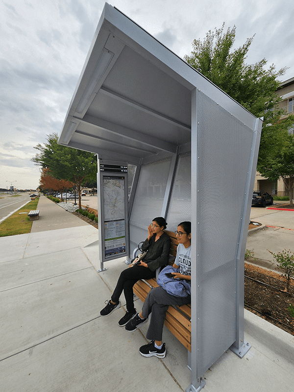 Signa Custom bus shelter has two passenger awaiting their bus while sitting on the bench inside the shelter