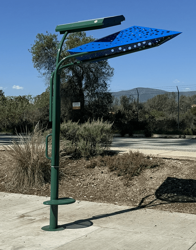 A signa custom bus shelter is seen from the right size with a green post and a blue leaf shaped top to provide shade to the single seat.