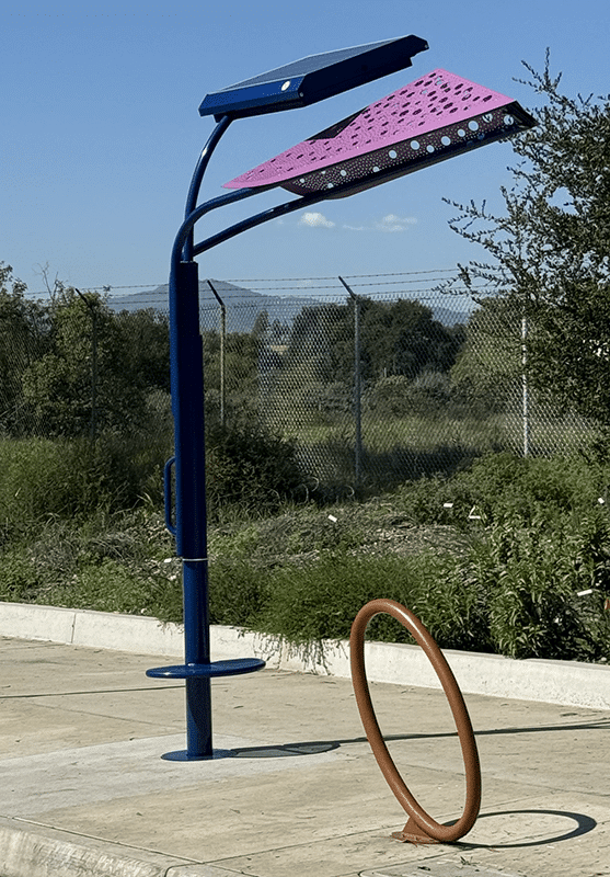 A signa custom bus shelter is seen from the right side with a blue post and a leaf shaped purple top and a round bike lock stand nearby.