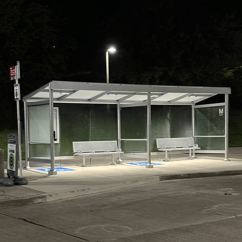 A Signa Empire bus shelter is seen at night lit up with two benches for seating and handicap marked sections with rider display.