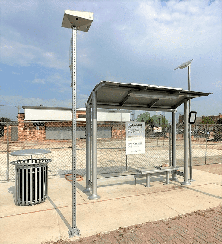 Right side view of a bus shelter with glass siding with an empty lot in the background and trash can close by