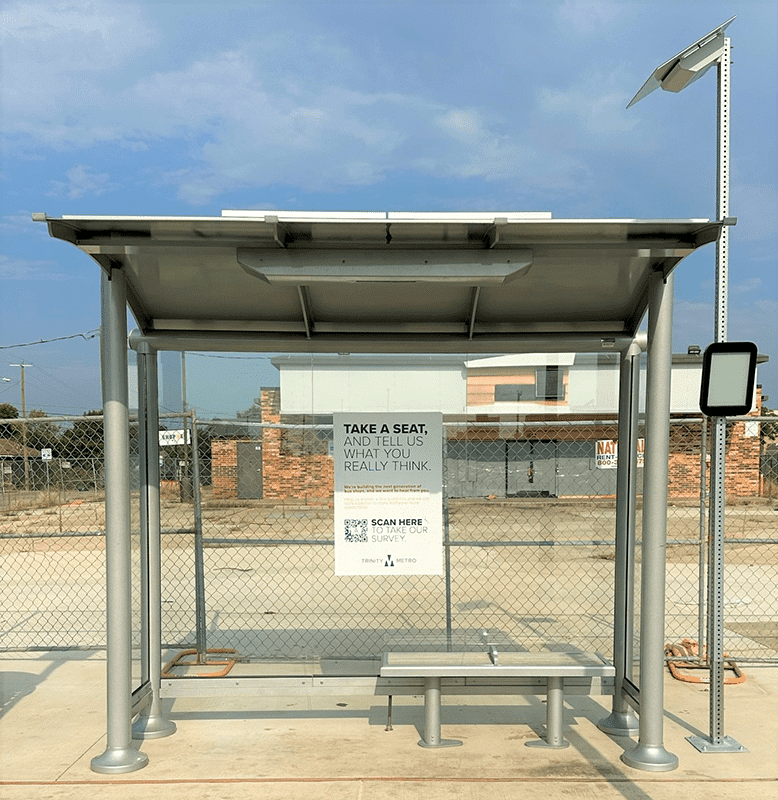 A head on view of a bush shelter with glass siding and a bench. In the background is a chain-link fence for an empty lot.