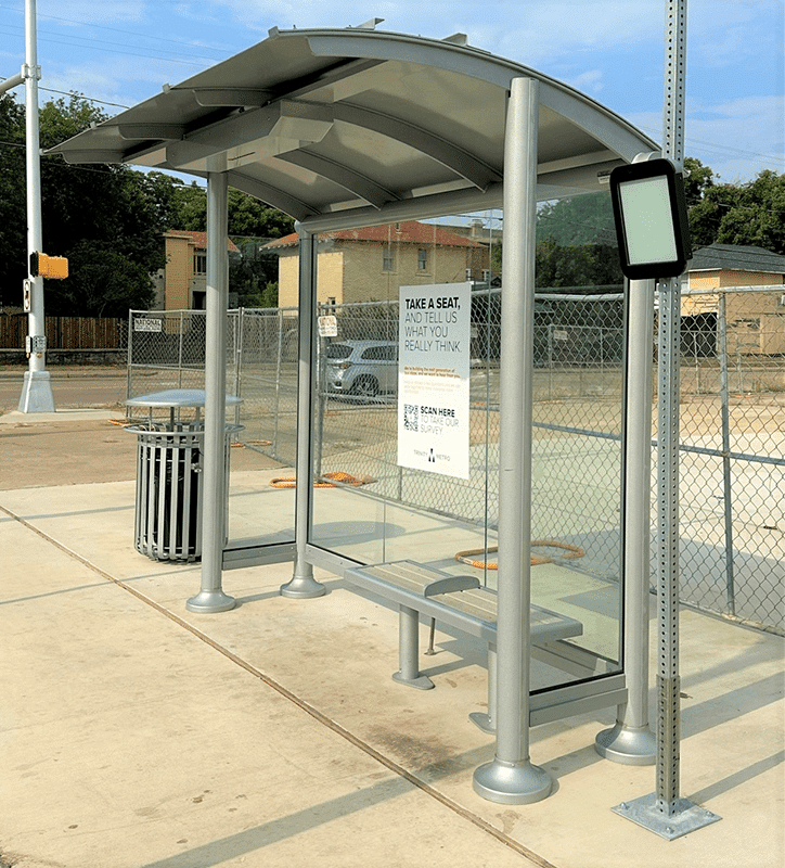 Left side view of a bush shelter with glass sides, a single bench, advertising on the back wall and a trash receptacle near by