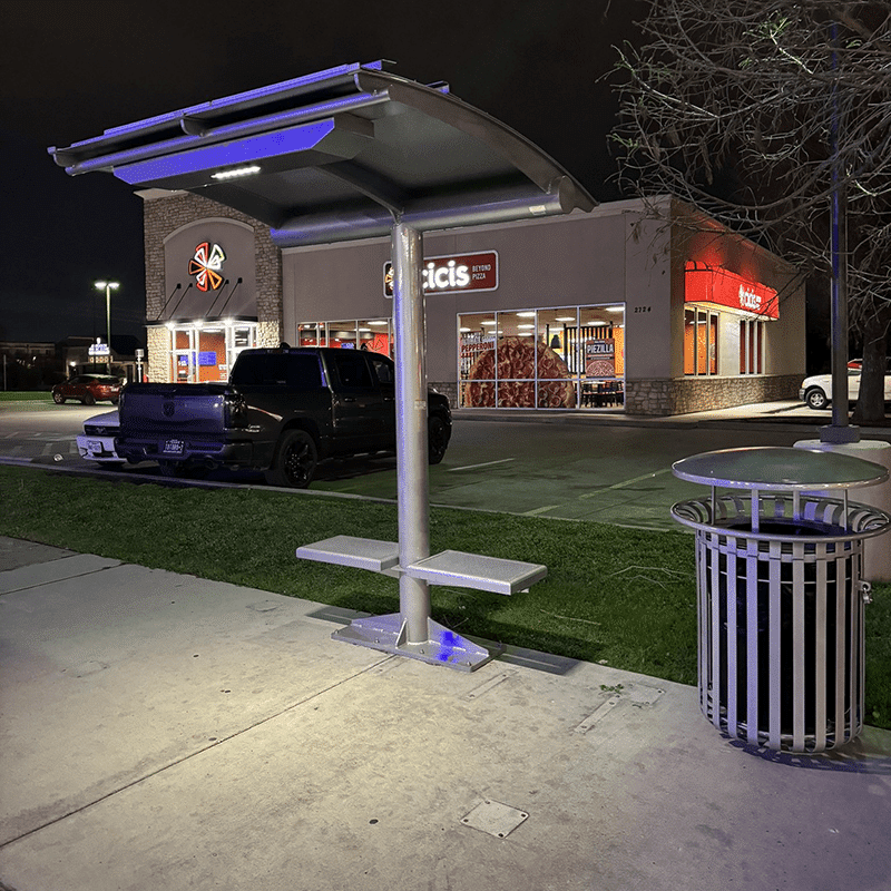 An Ecoshade is seen at night in front of a parking lot with two seats, a trash receptacle and ample lighting.