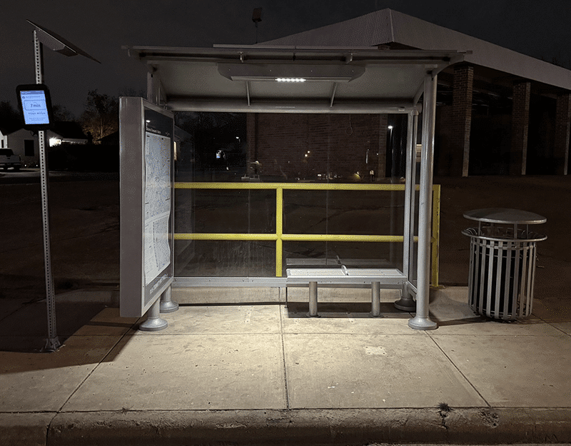 A head on view of a lit bus shelter at night with glass sides, advertising double sided display and a bench