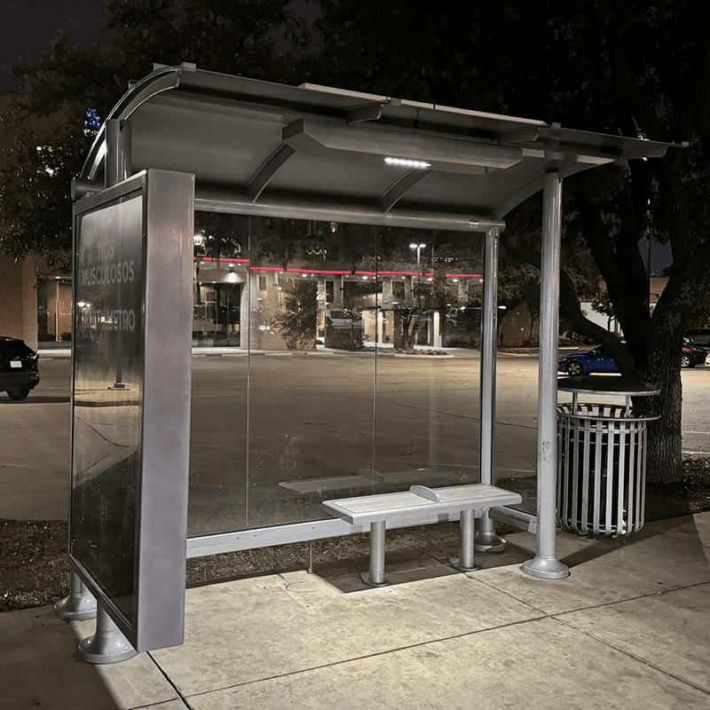 Right side view of a lit bus shelter at night with an advertising display, bench and trash receptacle