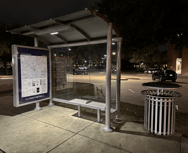 Left side view of a lit bus shelter with a rider information displayed on the right side and a trash can to the left of the shelter