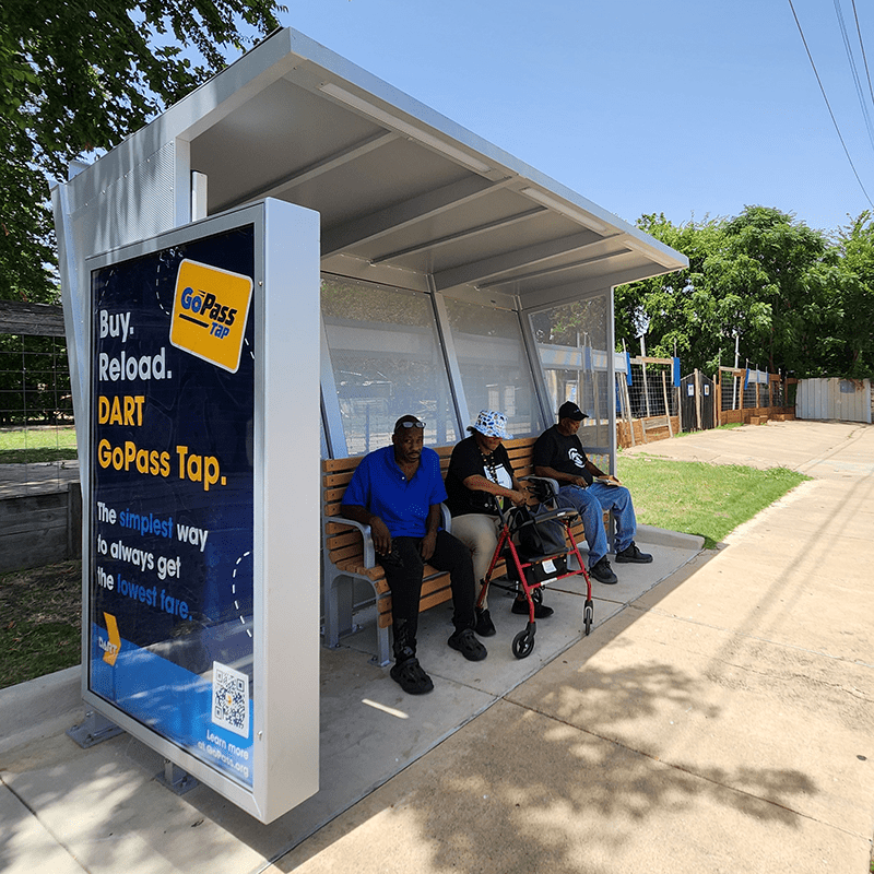A Siga Custom bus shelter seen from the right side displays a DART advertisement for three passengers sitting on the shelter's bench