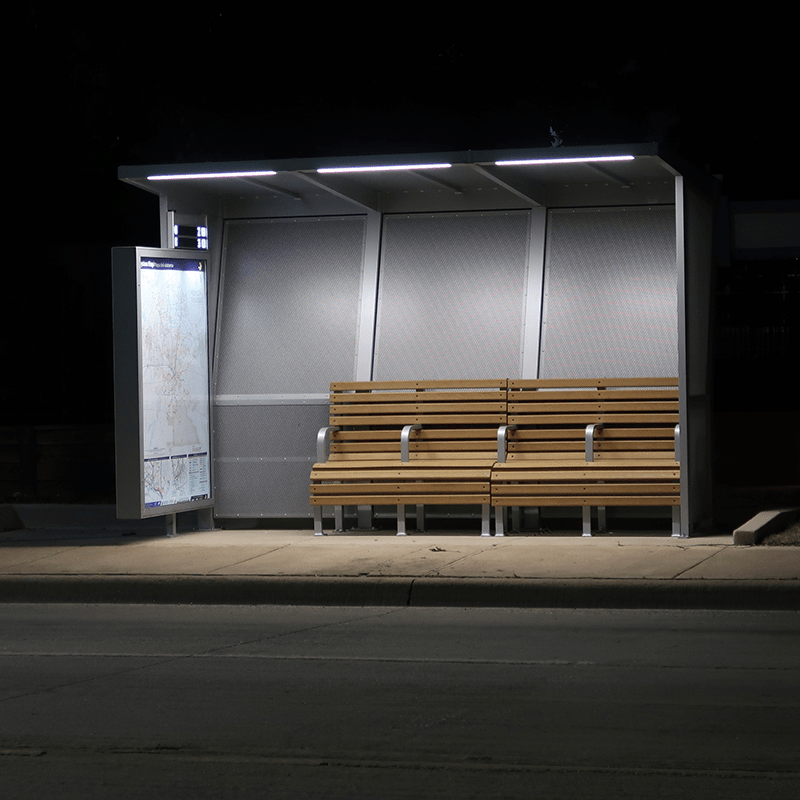 A Siga Custom bus shelter at night is lit displaying two benches and rider information display