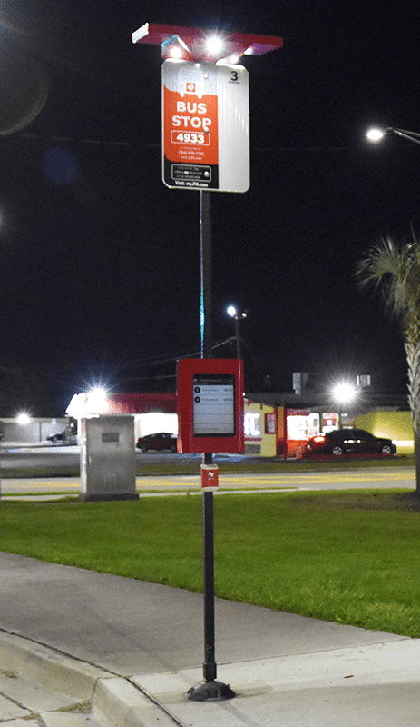 A front left side view of a bus stop post with information display for riders to see when the next bus will arrive and a lit up station number sign on top.