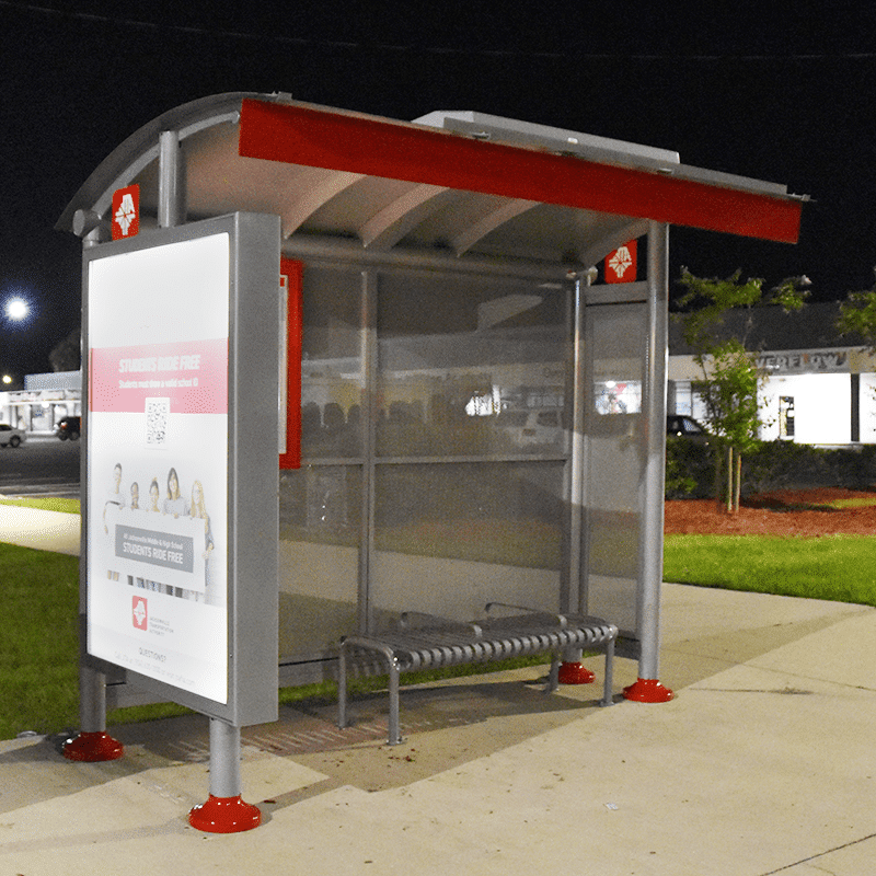 A bus shelter is seen at dusk with an advertising left side display and bench seating for three.
