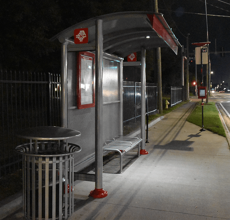 A signa sunset is seen from the left side at night with a trash receptacle on the left, rider display inside the shelter and a bench for three, all well lit.