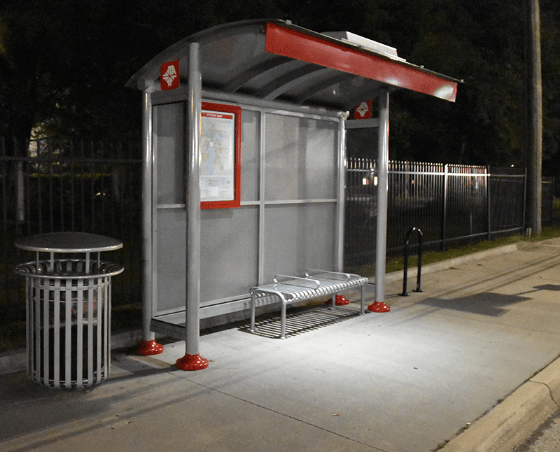 A signa sunset bus shelter is seen from the front left with a trash receptacle on the left outside and rider display and bench inside and a bike stand to the right of the shelter.