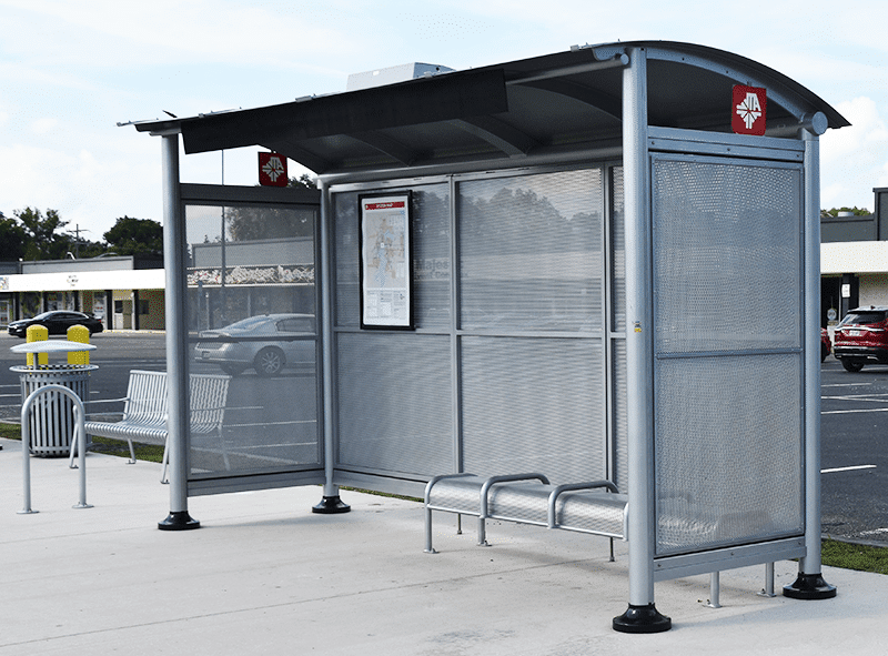 A signa sunset bus shelter is seen from the front right with a trash receptacle with bench seating on the outside left and inside the shelter is a rider information display and bench seating for four.