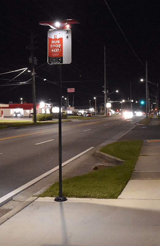 A bus stop is seen at night with a bus stop post with rider signage and a light on top for drivers to see riders waiting.