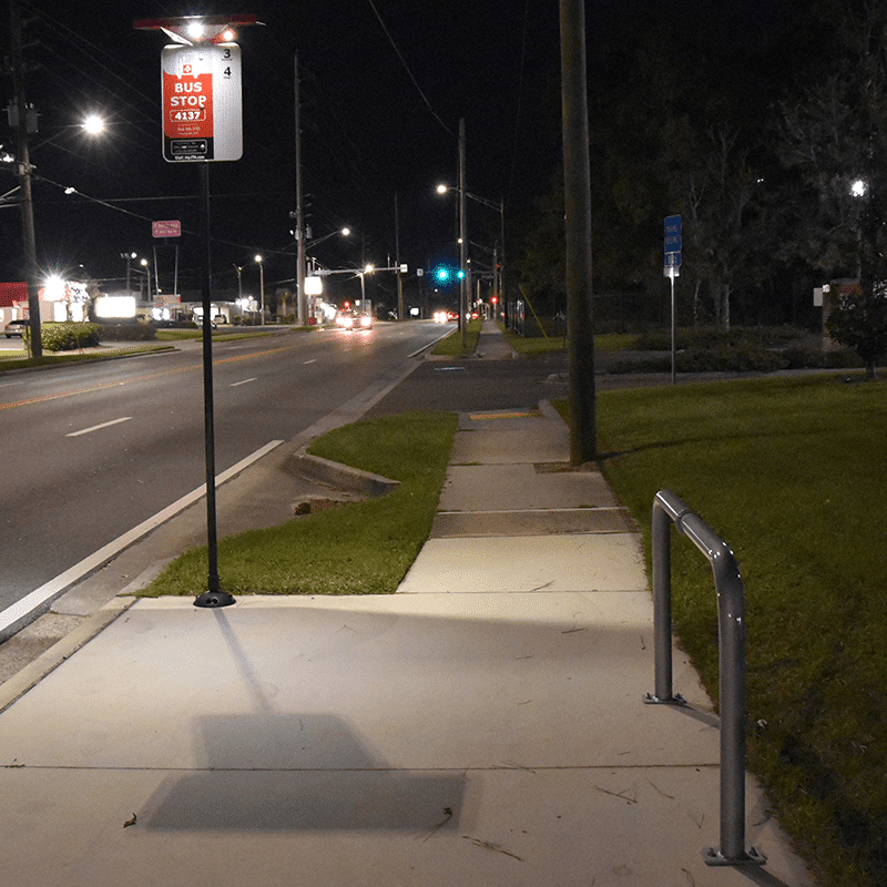 A bus stop is seen at night with bus signage and a light on the bus stop post located next to the street and a bike stand to the right of the sidewalk.