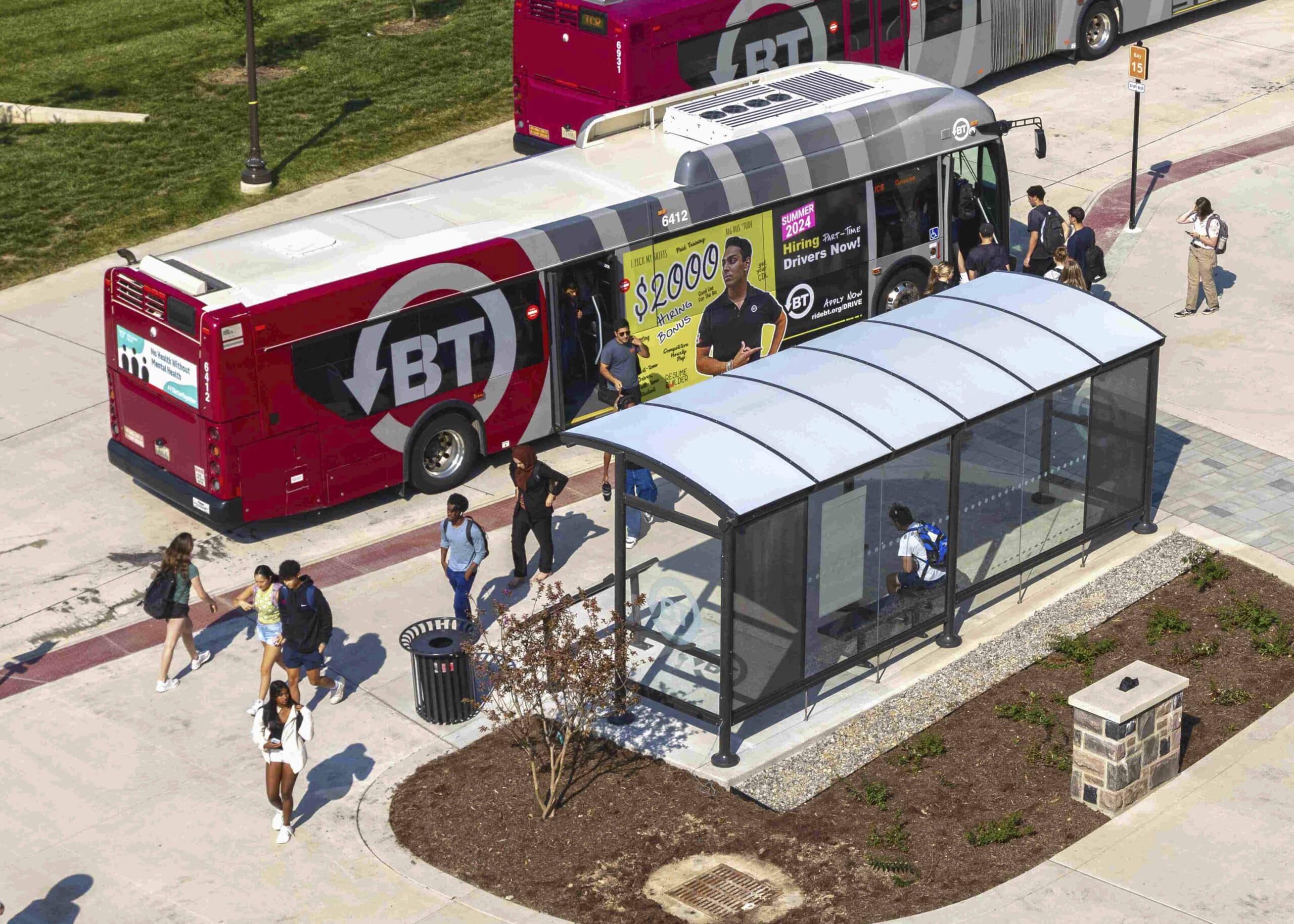 A bus shelter is seen from above with a view of the back left of the shelter with a trash receptacle on the left and back glass paneling so you can see the rider sitting on a bench inside the shelter as they await a bus.