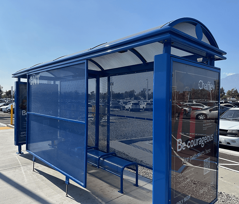 A blue bus shelter is seen from the front right with three full sides and the fourth with one center panel leaving entrance on both sides for seating inside the shelter for three.