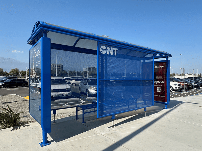 A bus shelter is seen from the front left for travelers at Ontario airport with an advertising display on both side panels and bench seating for three inside.