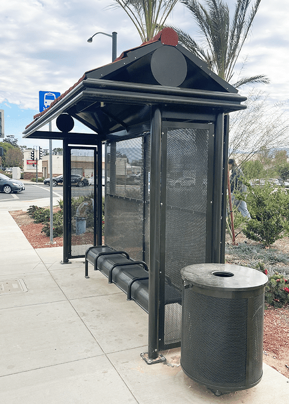 A bus shelter is seen from the front right with a trash receptacle on the outside right side and bench seating for four inside the shelter.