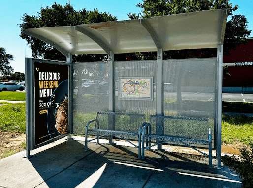 A siga custom bus shelter is seen from the front with a advertising display panel on the left and two benches under the shelter off to the right.
