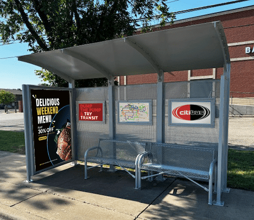 A siga custom bus shelter is seen from the front with an advertisement display on the left side panel, three back advertisement displays on the back and two benches.