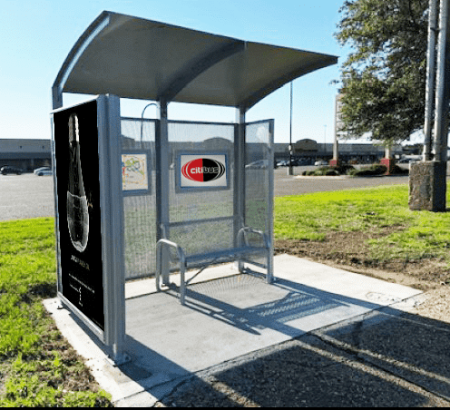 A siga custom bus shelter is seen from the front left with an advertisement display on the left side panel and two interior advertisement displays and one bench.