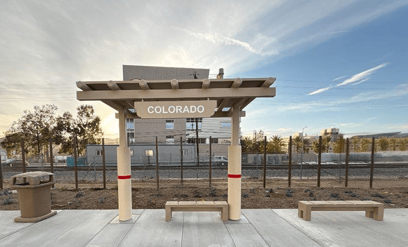 A bus shelter is seen from the front with signage reading Colorado, bench seating under the shelter and one to the right and a trash receptacle on the left.