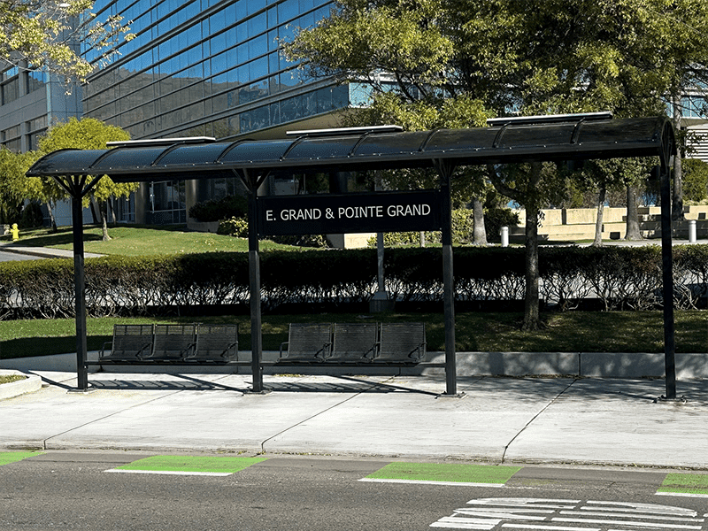 A signa voyager bus shelter is seen from the front with signage reading E. Grand & Pointe Grand with six seats under the shelter.