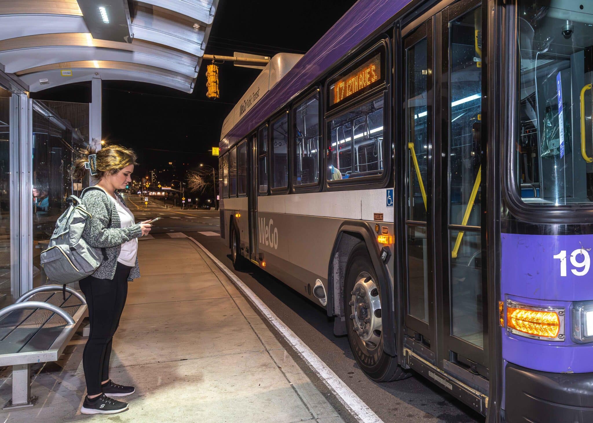 A woman is seen standing under a bus shelter as a bus pulls up to pick her up at night.