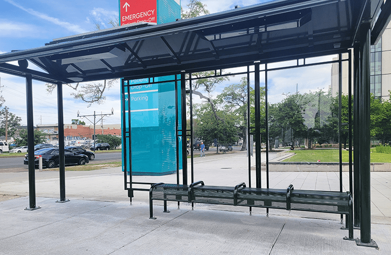 A bus shelter is seen from the front with two back clear glass panels and one blue glass panel and a bench with seating for four.